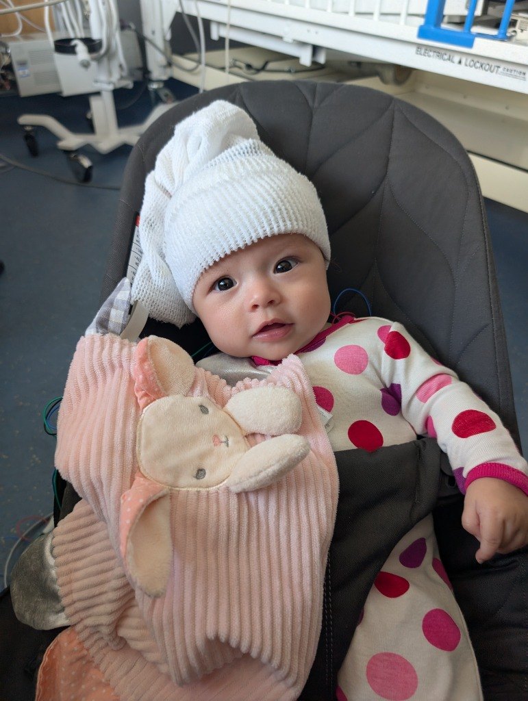 Baby Jojo in polka-dot pajamas and a white EEG cap, holding a pink bunny and looking at the camera