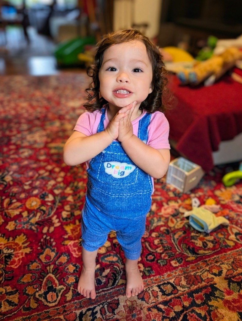 Jojo as a toddler, standing on a rug at home in blue overalls, smiling with her hands clasped