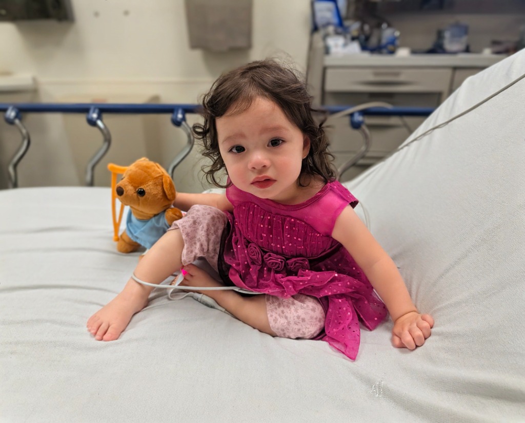 Jojo as a toddler in a pink party dress, sitting on a hospital bed with a monitor wire on her toe and a teddy bear beside her