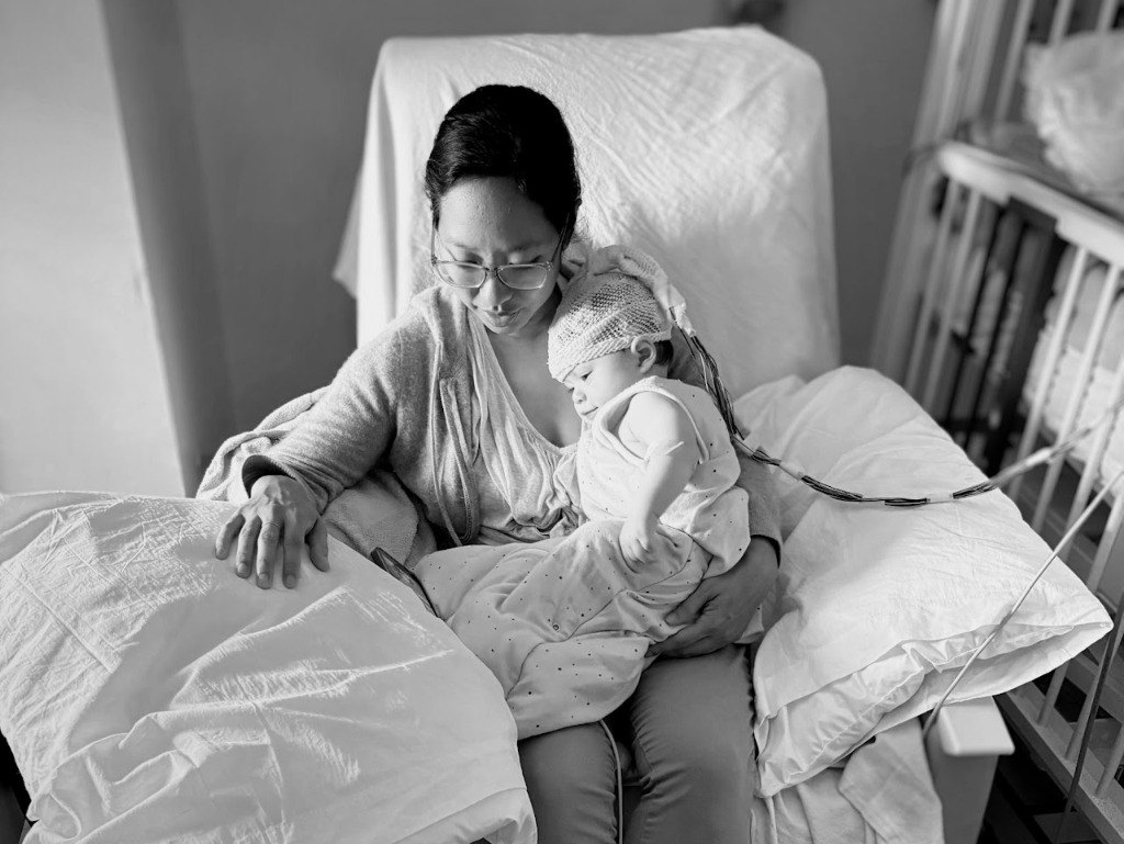 Black and white photo of Jojo's mom holding her in a hospital chair while Jojo wears an EEG head wrap